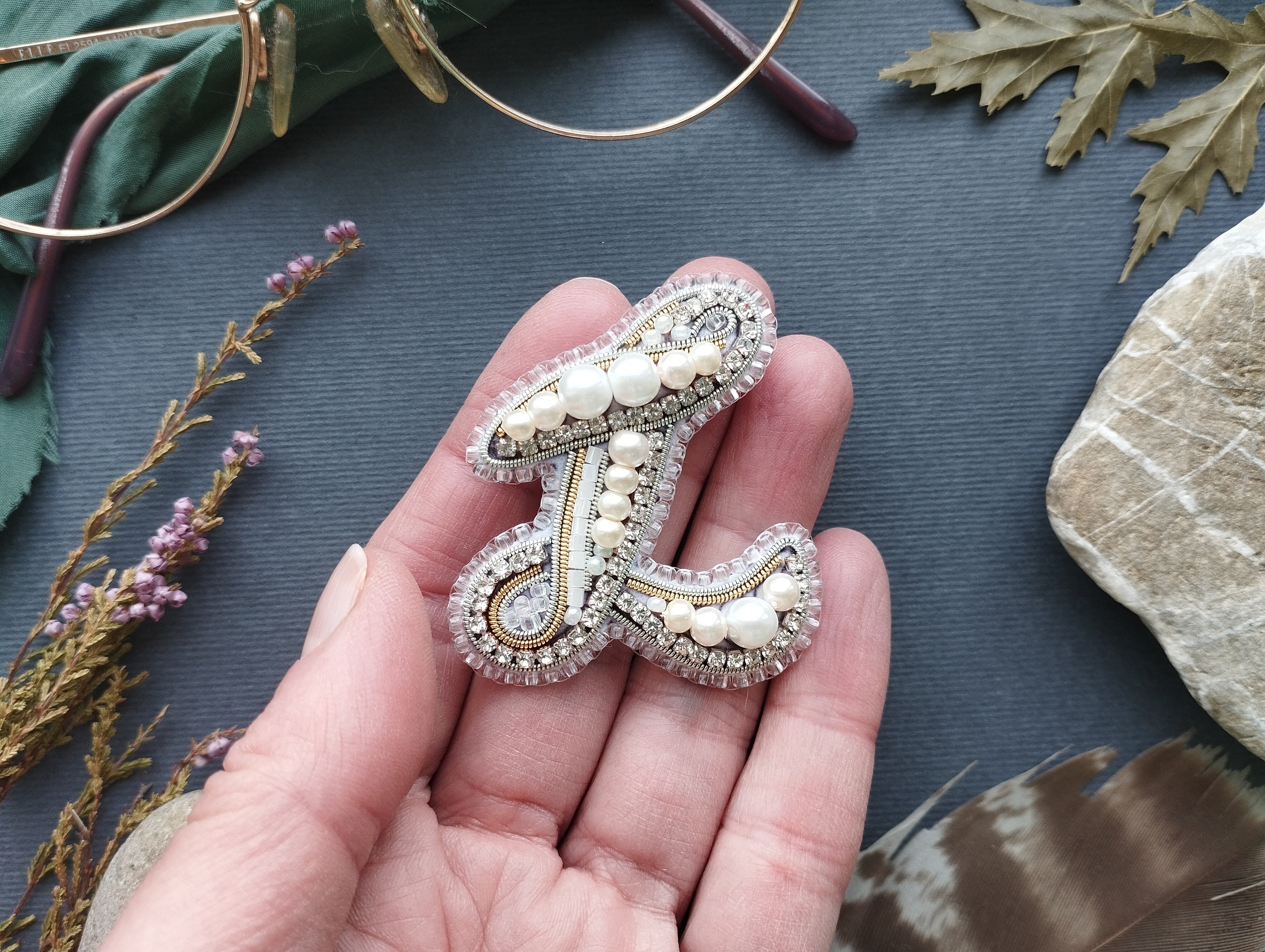 A hand holding a decorative letter 'L' made of beads and pearls against a dark background.